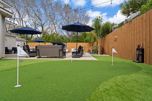 Backyard with artificial turf, patio seating under blue umbrellas, putting green, and wooden fence