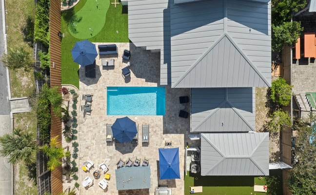 Aerial view of modern home with pool, patio seating, blue umbrellas, manicured lawn, and metal-roof structures