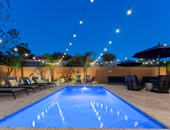 Illuminated backyard pool at dusk with lounge chairs, umbrellas, string lights, and wooden fence