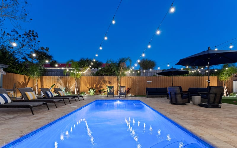 Illuminated backyard pool at dusk with lounge chairs, string lights, wooden fence, and black umbrellas